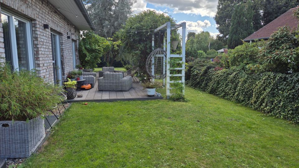 Eine Terrasse mit Sitzmöbeln aus Rattan neben einem Haus mit Klinkerfassade, umgeben von einem gepflegten Garten mit Grasfläche, Hecken und Bäumen. Im Hintergrund ist ein Holzgerüst mit einem Hängesessel angebracht, und der Himmel ist teilweise bewölkt.