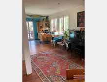 Wohnraum mit Holzboden und großen Fenstern, durch die Tageslicht einfällt. Im Vordergrund liegt ein gemusterter Teppich, rechts steht ein schwarzes Klavier, und im Hintergrund befindet sich eine offene Küche mit Holzmöbeln und Blick auf eine Terrasse.