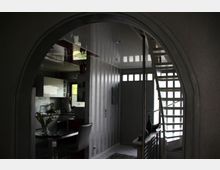 Blick durch einen Rundbogen auf den Innenraum eines modernen Hauses mit grauen Wänden. Links befindet sich eine Küche mit weißen Hängeschränken und einer Arbeitsfläche, rechts eine Treppe mit offenen Stufen und lichtdurchlässigen Glasbausteinen im Hintergrund.