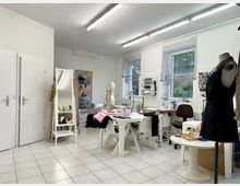 Ein heller Arbeitsraum mit Nähutensilien, mehreren Tischen und Nähmaschinen. An den Wänden befinden sich Regale mit Garnrollen, ein großer Spiegel steht neben weißen Türen, und eine Schneiderpuppe mit einem schwarzen Kleid ist sichtbar.