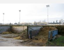 Kompostbereich mit Holzwänden und einer blauen Metallmulde, gefüllt mit organischen Abfällen und Stroh. Im Hintergrund sind ein Sportstadion mit Tribünen, Flutlichtmasten und umliegende Gebäude sichtbar.