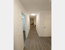 Langer, neutral beleuchteter Flur in einer Wohnung mit Holzoptik-Boden, weißen Wänden und mehreren Türen; am Ende des Flurs ist ein Essbereich mit Tisch und Stühlen sichtbar.