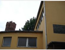 Gelbes Gebäude mit rechteckigen Fenstern im Vordergrund, daneben ein gemauerter, runder Turm aus rotem Backstein mit Zinnen; im Hintergrund ein Baum vor bewölktem Himmel.