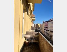 Ein schmaler Balkon eines historischen Mehrfamilienhauses mit gelber Fassade und dekorativen Stuckelementen. Der Balkon ist mit einem Boden aus Holzfliesen, zwei Holzstühlen und einer Sichtschutzmatte aus Naturmaterialien ausgestattet; im Hintergrund sind weitere Altbauten sowie eine Straße unter klarem, blauem Himmel zu sehen.