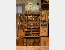 Eine alte Werkstatt mit einem offenen Holzschrank, der eine Vielzahl von Handwerkzeugen wie Hobel, Meißel, Sägen, Schraubenzieher und Scheren zeigt. Im Hintergrund sind eine Wanduhr und eine Fensterfront zu sehen, die von einer schlichten, verwitterten Wand umgeben sind.