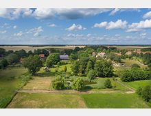 Luftaufnahme einer ländlichen Umgebung mit Wiesen, einem Teich und von Bäumen gesäumten Bereichen. Im Hintergrund stehen mehrere Gebäude mit roten Dächern, umgeben von Feldern und Wäldern unter einem leicht bewölkten Himmel.