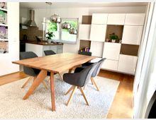 Esszimmer mit einem großen Holztisch und sechs grauen Polsterstühlen auf einem hellen Teppich. Im Hintergrund befinden sich eine moderne weiße Einbauküche, ein großes Regal mit weißen Fronten und Pflanzen sowie ein Fenster, das Tageslicht hereinlässt.