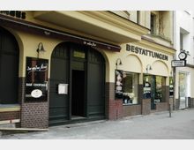 Fassade eines Gebäudes mit zwei nebeneinanderliegenden Geschäften: links eine Bar mit der Aufschrift „Zur weißen Maus – Bar-Cocktails“, rechts ein Bestattungsinstitut mit dem großen Schriftzug „Bestattungen“ über der Tür. Die Fassade ist beige mit braunen Klinker-Elementen, und beide Eingänge sind von gewölbten Bögen umrahmt.