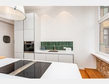 Moderne, minimalistische Küche mit weißen, griffarmen Schränken, eingebautem Backofen und einer Arbeitszeile mit Spüle vor einer grünen Fliesenrückwand. Im Vordergrund steht eine Kochinsel mit Induktionskochfeld; rechts sorgt ein großes Fenster mit gemauerter Brüstung und Holzstufen für Tageslicht.