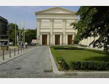 Das Bild zeigt die Fassade des Maxim-Gorki-Theaters in Berlin, ein klassizistisches Gebäude mit Säulen und Reliefs über den drei großen Eingangstüren. Im Vordergrund sind ein gepflasterter Weg, Grünflächen und einige Fußgänger zu sehen.