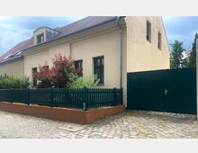 Ein beige gestrichenes Einfamilienhaus mit Satteldach und dunkelgrünen Fensterrahmen, umgeben von einem kleinen Garten mit Ziersträuchern und einem dunkelgrünen Lattenzaun. Vor dem Haus liegt ein gepflasterter Weg, und rechts befindet sich ein großes, passendes Gartentor.