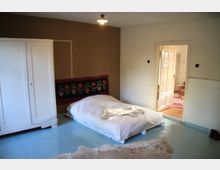 Schlafzimmer mit einfachem, niedrigen Bett mit weißer Bettwäsche auf hellem Holzboden. Im Raum befinden sich ein weißer Schrank, eine bunte Wanddekoration und eine offene Tür, die zu einem angrenzenden, teilweise sichtbaren Raum führt.