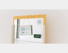 Ein rechteckiger Spiegel mit einer Sperrholz-Umrandung, an dem ein Schild mit der Aufschrift „Photoshoot! Do Not Disturb. Please Use Other Door“ hängt. Im Spiegel ist ein heller Raum sowie ein grünes Sofa im Hintergrund zu sehen.