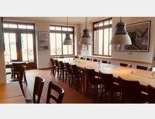 Heller Gastraum mit großen Fenstern, einem langen Holztisch mit mehreren Stühlen und drei großen, hängenden Industrielampen. An den Wänden hängen Fotos, und durch die Fenster ist eine Terrasse sichtbar.