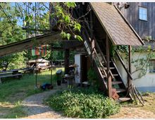 Außenbereich eines rustikalen Gebäudes mit Holztreppe, die zu einem überdachten Eingang führt. Im Vordergrund ein mit Kopfsteinpflaster gestalteter Weg, umgeben von Pflanzen; im Hintergrund Holztische und ein schattiger Garten mit Bäumen.