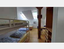 Ein einfach eingerichtetes Schlafzimmer mit einem Etagenbett, zwei Einzelbetten, einem Holztisch mit blauen Stühlen und Regalen. Der Raum hat zwei Dachfenster, eine sichtbare Holzstütze in der Mitte und einen hellen Bodenbelag.