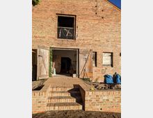 Backsteingebäude mit offenstehenden hölzernen Toren und einer Eingangstreppe aus Ziegelsteinen. Das Gebäude zeigt Spuren von Renovierungsarbeiten, mit blauen Säcken und Baumaterialien im Eingangsbereich.