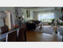 Wohn- und Essbereich mit Esstisch und gepolsterten Stühlen im Vordergrund, dahinter Sofa und Couchtisch auf einem großen Teppich. Ein breites Fenster mit transparenten Vorhängen und vielen Zimmerpflanzen sorgt für Tageslicht; rechts stehen Glasvitrinen und Dekoration, links führt ein Rundbogen in den Flur.