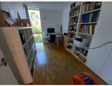 Ein heller Arbeitsraum mit Parkettboden, ausgestattet mit weißen Bücherregalen voller Bücher und Ordner. Im Hintergrund steht ein Schreibtisch mit einem Monitor und Bürostuhl, daneben ein großes Fenster, das auf grüne Bäume blickt.