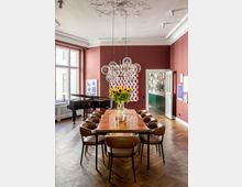 Ein stilvoll eingerichteter Raum mit holzgetäfeltem Boden, einem langen Esstisch aus Holz und braunen Stühlen. Im Hintergrund befinden sich eine Glasvase mit Sonnenblumen, ein Flügel, Wandkunst mit geometrischen Mustern und eine aufwendig verzierte Deckenleuchte mit Glaslampen.