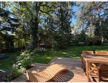 Holzterrasse mit Tisch, Stühlen und geschwungener Liege vor einem großen, gepflegten Garten mit Rasenfläche. Im Hintergrund stehen hohe Laub- und Nadelbäume sowie dichtes Strauchwerk, teilweise mit Blick auf ein Hausdach durch die Bäume.
