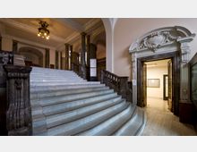 Großes Treppenhaus in einem historischen Gebäude mit weißen Marmorstufen, verzierten dunklen Geländern und Wandpfeilern aus poliertem Stein. Rechts neben der Treppe befindet sich ein aufwendig gestaltetes Portal mit Stuckdekorationen, das in einen beleuchteten Raum führt.