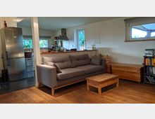 Wohnzimmer mit grauem Sofa, kleinem Holztisch und Holzboden, im Hintergrund eine offene Küche mit Edelstahlkühlschrank, Dunstabzugshaube und Fenstern mit Blick ins Grüne. Rechts ein Bücherregal und Holzmöbel.