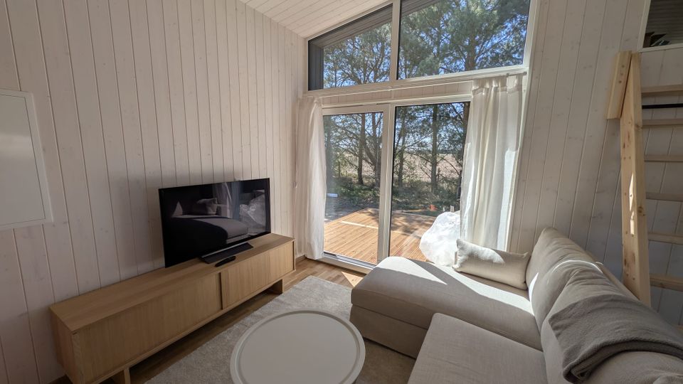 Heller Wohnbereich in einem Holzhaus mit weiß vertäfelten Wänden und schräger Decke, Sofa, Couchtisch und TV auf Lowboard. Große bodentiefe Fensterfront mit Oberlicht und weißen Vorhängen führt auf eine Holzterrasse mit Blick auf Bäume.