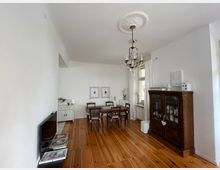 Esszimmer mit Holzdielenboden, weiß gestrichenen Wänden und einem verzierten Kronleuchter an der Decke. Ein Holztisch mit Stühlen steht im Zentrum, ein antiker Glasschrank und weiße Vorhänge umrahmen die Fenster auf der rechten Seite, während links ein kleiner weißer Beistellschrank und ein Fernseher sichtbar sind.