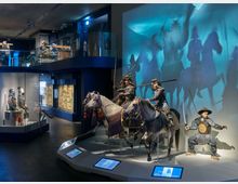 Ausstellungsraum eines Museums mit dem Schwerpunkt auf japanischer Geschichte, dargestellt durch samuraiähnliche Figuren in traditioneller Rüstung, teils auf Pferden. Im Hintergrund sind beleuchtete Schattenprojektionen von Reiterszenen zu sehen, und Glasvitrinen mit weiteren Artefakten wie Helmen und Waffen säumen den Raum.