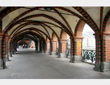 Überdachter Gang mit gemauerten Backsteinbögen und verzierten Eisenbrüstungen, Teil der Oberbaumbrücke in Berlin; im Hintergrund sind Gebäude und der Fluss Spree zu sehen.