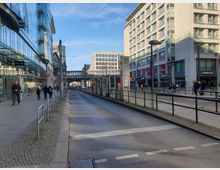 Straßenansicht mit Straßenbahnhaltestelle, flankiert von modernen Glas- und Betongebäuden. Links ein Einkaufszentrum mit Glasfassade, rechts ein Gebäude mit Gastronomie und Geschäften im Erdgeschoss; im Hintergrund eine Brücke mit Bahnstrecke.