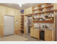 Ein minimalistischer Raum mit Sperrholzverkleidungen an den Wänden und offenen Regalen, die Küchenutensilien, Geschirr und Lebensmittel zeigen. Eine kleine Küchenzeile mit Spüle, Wasserkocher und weiteren Geräten sowie eine graue Tür und eingepasste Holzstufen prägen den Raum, der eine funktionale Einrichtung bietet.