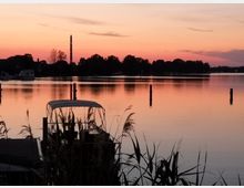 Der Sonnenuntergang spiegelt sich auf einem ruhigen See, im Vordergrund sind Schilfgräser und ein kleiner Bootssteg mit einem überdachten Boot zu sehen. Im Hintergrund ragen Bäume und ein beleuchteter Schornstein vor dem orange-rosa Himmel hervor.