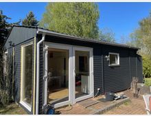 Kleines, dunkel verkleidetes Holzhaus im Garten mit flachem, leicht geneigtem Dach, Dachrinne und großen Glas-Doppeltüren sowie Seitenfenster. Vor dem Eingang liegt eine gepflasterte Terrasse; im Hintergrund stehen hohe Bäume unter blauem Himmel.