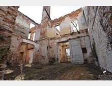 Innenhof eines verfallenen Backsteingebäudes ohne Dach, mit freiliegenden Ziegelwänden, großen leeren Fensteröffnungen und einem gemauerten Bogen. Im Vordergrund liegen überwucherte Bodenflächen und eine verwitterte Tür in einem teilweise erhaltenen Innenraum.