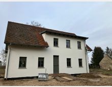 Zweistöckiges Einfamilienhaus mit weißer Fassade und rotem Ziegeldach in ländlicher Umgebung. Vor dem Haus liegt unbefestigter Boden mit Baumaterialien, im Hintergrund sind Bäume und ein weiteres Gebäude sichtbar.