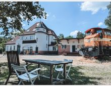 Ein historisches Gebäude mit weißer Fassade, roten Ziegeldetails und einem markanten Turm, daneben eine orangefarbene alte Lokomotive. Im Vordergrund stehen Gartenmöbel auf einer Wiese, umgeben von Bäumen bei sonnigem Wetter.