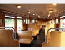 Innenraum eines historischen Eisenbahnwaggons mit hölzernen Sitzbänken, Metallgriffen und großen Fenstern. Oberhalb der Sitze befinden sich Gepäckablagen, und außen ist eine grüne Landschaft zu sehen.