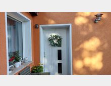 Eingangsbereich eines Hauses mit orangefarbener Putzfassade, weiß gerahmter Haustür mit schmalen Glasfenstern und dekorativem Türkranz. Daneben ein Fenster mit breiter Fensterbank und mehreren Blumentöpfen sowie eine Außenwandleuchte.