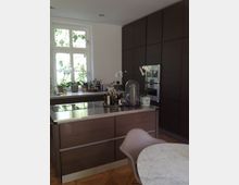 Moderne Küche mit dunklen Holzschränken, einer Kücheninsel mit Edelstahloberfläche und einem runden Marmortisch im Vordergrund. Ein großes Fenster im Hintergrund lässt Tageslicht herein, daneben sind Pflanzen und Gewürzflaschen auf der Fensterbank sichtbar.