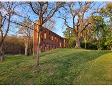 Backsteinruine einer historischen Kirche auf einem grasbewachsenen Hügel, umgeben von hohen Bäumen im Abendlicht; im Hintergrund dichter Wald.