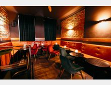 Gemütlicher Lounge- oder Barbereich mit langer, gepolsterter Leder-Sitzbank entlang der Wände, kleinen runden Tischen und gepolsterten Stühlen. Sichtbare Ziegelwände, dunkle Wandflächen mit Wandleuchten und schwere Vorhänge prägen die gedämpfte Innenraum-Atmosphäre.