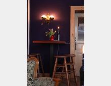 Ein kleines Café-Interieur mit einer dunkellila Wand, an der eine Wandlampe mit zwei leuchtenden Glühbirnen befestigt ist. Darunter steht ein schmaler Holztisch mit Barhockern und einer roten Vase mit Tulpen, sowie einer brennenden Kerze auf einer Glasflasche. Rechts daneben befindet sich eine Tür mit einem hölzernen Rahmen.