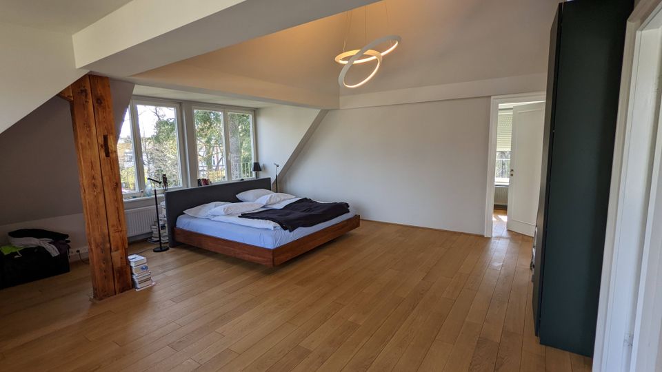 Geräumiges Schlafzimmer im Dachgeschoss mit Holzfußboden, schrägen Deckenflächen und sichtbarem Holzbalken. Ein niedriges Bett steht vor großen Fenstern mit Blick ins Grüne; an der Decke hängt eine moderne Ring-Pendelleuchte, rechts führt eine Tür in einen Nebenraum.