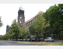 Straßenansicht eines großen historischen Gebäudes mit dunklem Steinturm und grünem Dach, flankiert von einer Reihe von Bäumen. Vor dem Gebäude befinden sich parkende Autos entlang einer Straße mit Mittelstreifen.