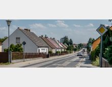 Straßenszene in einem Vorort mit einer Reihe von Einfamilienhäusern mit Satteldächern auf der linken Seite und einer schmalen Straße mit parkenden Autos. Im Hintergrund sind Bäume, ein Bushaltestellenschild und ein mehrstöckiges Gebäude sichtbar.