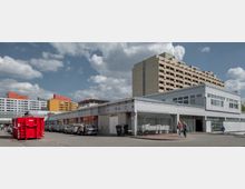 Eine städtische Szene mit einem flachen Gewerbegebäude im Vordergrund, Parkplätzen und einem roten Container. Im Hintergrund stehen mehrstöckige Wohngebäude in gelben, weißen und orangen Farben, unter einem wolkigen Himmel.