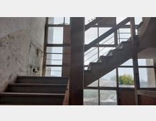 Treppe in einem verlassenen Gebäude mit verfallenen Wänden und großen Fensterfronten aus Glas, die den Blick auf eine bewachsene Außenlandschaft freigeben. Die Metallgeländer und die sichtbaren Abnutzungsspuren deuten auf einen vernachlässigten Zustand hin.