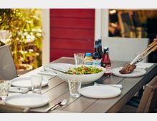 Ein gedeckter Gartentisch mit grauer Tischplatte, auf dem eine große Schüssel mit Salat, Teller, Gläser, Grillsoßen, Senf sowie Bierflaschen stehen; im Hintergrund eine rote Holzwand und Pflanzen. Eine Hand mit Grillzange legt ein Stück Fleisch auf einen Teller.